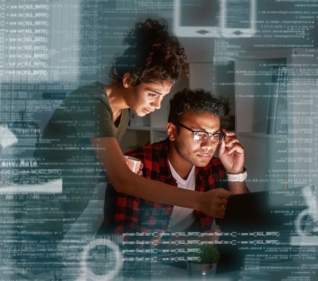 Two devs working together. One femal with brown shirt and curly hair and one male with red and black plaid checkered shirt and glasses.